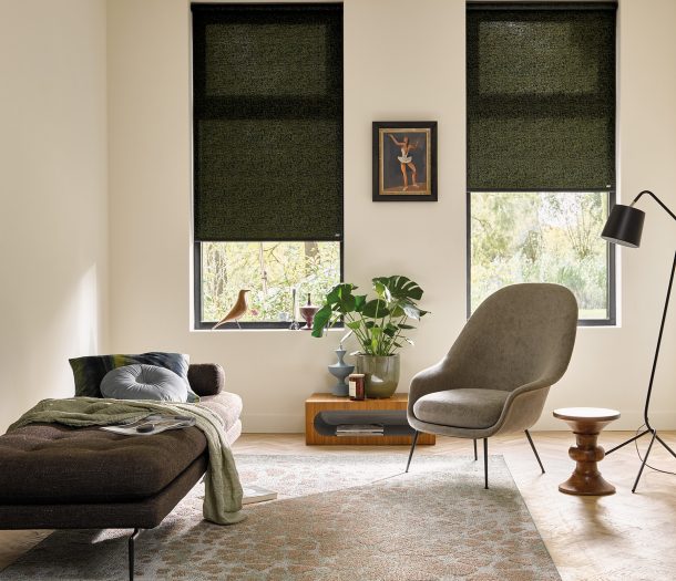 Woonkamer in natuurlijke stijl met vloerkleed met print, bankje en leesstoel, licht transparante rolgordijnen in donkergroen voor de hoge ramen.